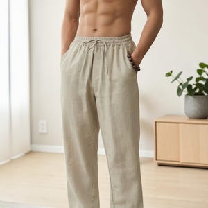May include: Beige trousers featuring an elastic waistband with a matching drawstring closure. These trousers have a relaxed, straight-leg fit and include side pockets. The fabric has a visible, textured weave consistent with linen or a similar material.