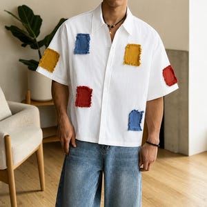 May include: A white short-sleeved button-down shirt featuring several rectangular patches with frayed edges in solid colors including mustard yellow, blue, and red, paired with loose-fitting blue denim jeans.