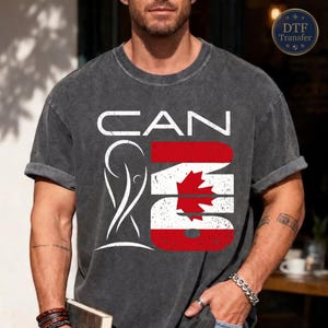 May include: A charcoal gray short-sleeve t-shirt featuring a white graphic that reads CAN with a stylized trophy icon and the number 26, where the number 26 is filled with the red and white Canadian flag design featuring a central maple leaf.