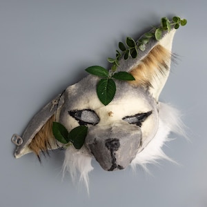 May include: A handcrafted animal mask featuring grey and white faux fur, pointed ears with brown accents, mesh eye coverings, and a dark nose. The mask is decorated with artificial green leaves and vines attached to the ears and forehead area.