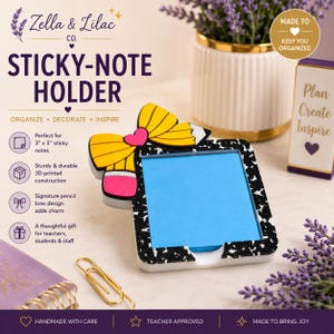 May include: A square sticky-note holder with a black and white composition notebook pattern, featuring a yellow pencil-shaped bow with a pink eraser and a small pink heart. The holder contains a stack of bright blue 3-inch by 3-inch sticky notes.