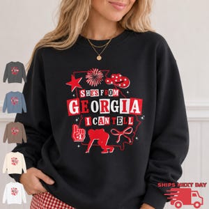May include: A black long-sleeve sweatshirt featuring a red and white graphic print that reads "SHE'S FROM GEORGIA I CAN TELL" with illustrations of a star, pom-pom, cowboy hat, foam finger, football player silhouette, and a bow.