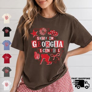 May include: A dark brown short-sleeve t-shirt featuring a red and white graphic print that reads "SHE'S FROM GEORGIA I CAN TELL" with icons including a star, firework, cowboy hat, foam finger, football player silhouette, and a ribbon bow.