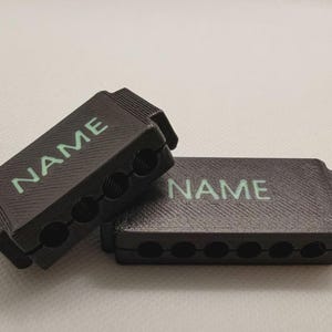 May include: Two rectangular black cable organizers with a textured surface and the word NAME printed in light green. One organizer features four circular cutouts along the side, while the other features six circular cutouts along the side.