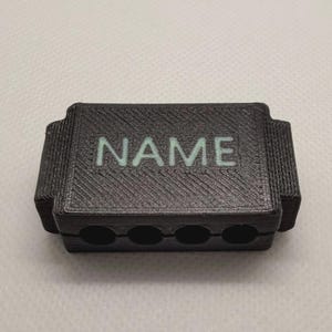May include: A rectangular black 3D printed object with a textured surface featuring the word NAME in light green capital letters. The object has four circular holes along the bottom edge and stepped side sections, showing a layered construction.