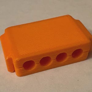 May include: A rectangular orange plastic block featuring four evenly spaced circular holes along the front edge. The object has a textured surface finish with visible horizontal lines and a split seam running horizontally across the front face.