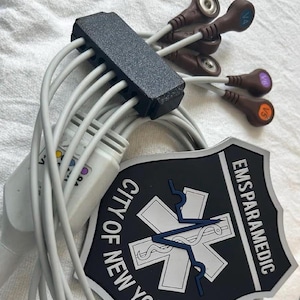 May include: A medical ECG cable set with gray wires, a black connector block, and brown electrode leads labeled V4, V5, and V6. A black and white shield-shaped patch features the text CITY OF NEW YORK EMSPARAMEDIC and a blue medical star symbol.