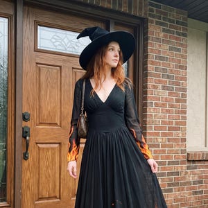 May include: A floor-length black dress with long sleeves featuring a vibrant orange and yellow flame print at the hem and cuffs. The dress has a V-neckline and a fitted bodice with a full, flowing skirt. A black pointed witch hat is worn on the head.