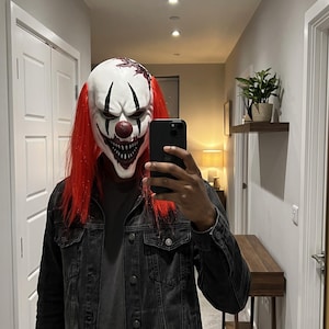 Peut inclure: Un masque de clown blanc avec un nez rouge, des marques noires autour des yeux et un large sourire denté. Le masque présente de longs cheveux rouge vif et un motif sombre et texturé sur le front, porté avec une veste en jean gris foncé.