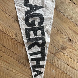以下が含まれることがあります： 大きな黒い太字のアルファベットで「LAGERTHA」とプリントされた白い三角形のペナント旗。生地は質感があり、わずかにシワが寄った外観で、先端を下に向けて垂直に配置されています。