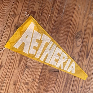 May include: A triangular yellow felt pennant featuring the word AETHERIA in large white block letters. The pennant has a thin white string loop attached to the top corner for hanging, with a textured fabric surface and visible stitching.