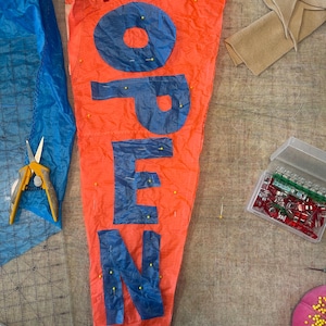 May include: A triangular orange fabric pennant featuring the word OPEN in large blue letters. The letters are secured to the orange fabric with yellow sewing pins. Red clips are attached to the top edge of the pennant during the assembly process.