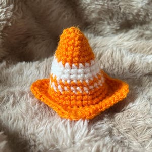 May include: A small, cone-shaped object crafted from orange and white yarn using a crochet technique. The item features a pointed top, a wide circular base, and alternating horizontal stripes of bright orange and white throughout the structure.