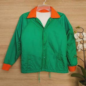 May include: A green windbreaker jacket featuring a bright orange collar and matching orange sleeve cuffs. The jacket has a white inner lining, a front snap-button closure, two side pockets, and a drawstring hem at the bottom for an adjustable fit.