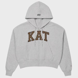May include: A light gray zip-up hoodie featuring a hood with the text GAP embroidered in black. The front displays the letters KAT in a large, collegiate-style font with a brown and black leopard print pattern and a black outline.