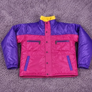 May include: A colour-blocked puffer jacket featuring a bright yellow collar, purple upper chest and sleeves, and a vibrant pink lower body. The jacket includes a central snap-button closure, two zippered chest pockets, and two lower side pockets.