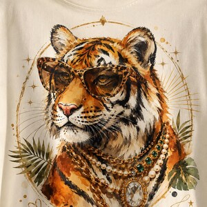 Könnte beinhalten: Ein cremefarbenes T-Shirt mit einer grafischen Illustration eines Tigers, der eine Sonnenbrille mit Leopardenmuster und geschichtete Goldketten mit Perlen und grünen Edelsteinen trägt, eingerahmt von einer goldenen Kreislinie und Palmblättern.