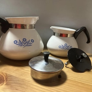 May include: Two white ceramic coffee pots with black handles and a blue floral pattern, featuring a metallic band around the neck. Included are a separate stainless steel lid with a black knob and a black circular component with a central metal pin.