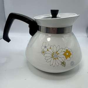 May include: A white enamel teapot featuring a floral pattern with yellow and white daisy-like flowers. The teapot has a black plastic handle, a matching black knob on the lid, and a silver-coloured metal band encircling the upper body section.