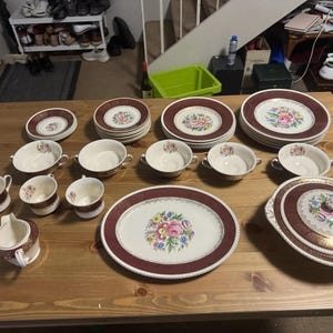 Può includere: Un set di stoviglie in ceramica con base color crema, bordo marrone scuro e un motivo floreale centrale. Il set comprende vari piatti, ciotole con manici, tazze da tè, una lattiera, un piatto da portata e una zuppiera con coperchio.