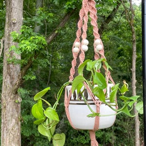 May include: A dusty rose macrame plant hanger featuring twisted cord patterns and round wooden beads, suspending a white ceramic pot with a black horizontal stripe design containing a trailing green pothos plant with heart-shaped leaves.