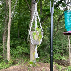Op de afbeelding: Een witte macramé plantenhanger met gedraaide koordpatronen en houten kralen, waaraan een gespikkelde grijze pot hangt met een hartbladige philodendronplant met groen en lichtgeel gevlekte bladeren en een witte kwast.