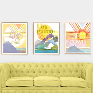 May include: Three watercolor prints with a mountain and sun design. The prints have the words "SLO chill", "SLO beautiful", and "SLO down" on them.