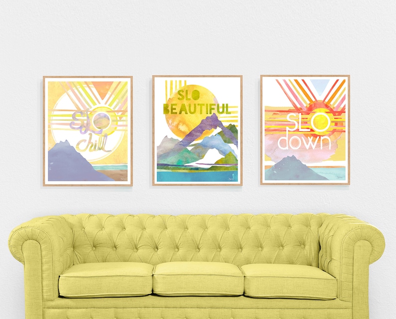 May include: Three watercolor prints with a mountain and sun design. The prints have the words "SLO chill", "SLO beautiful", and "SLO down" on them.