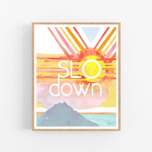 May include: A watercolor painting of a sunset with the words "slow down" in white text. The painting features a yellow sun with red and orange rays, a pink and blue sky, and a blue mountain in the foreground.