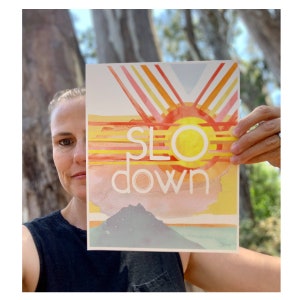 May include: A watercolor print with a sunset scene and the words "SLO down" in white text. The print features a yellow sun, orange and pink clouds, and a mountain range in the background.