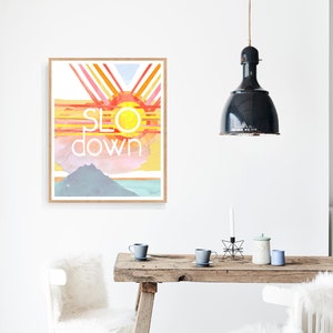 May include: A watercolor print with a sunset scene and the words "slow down" in white text. The print is framed in a light wood frame and is hanging on a white wall.