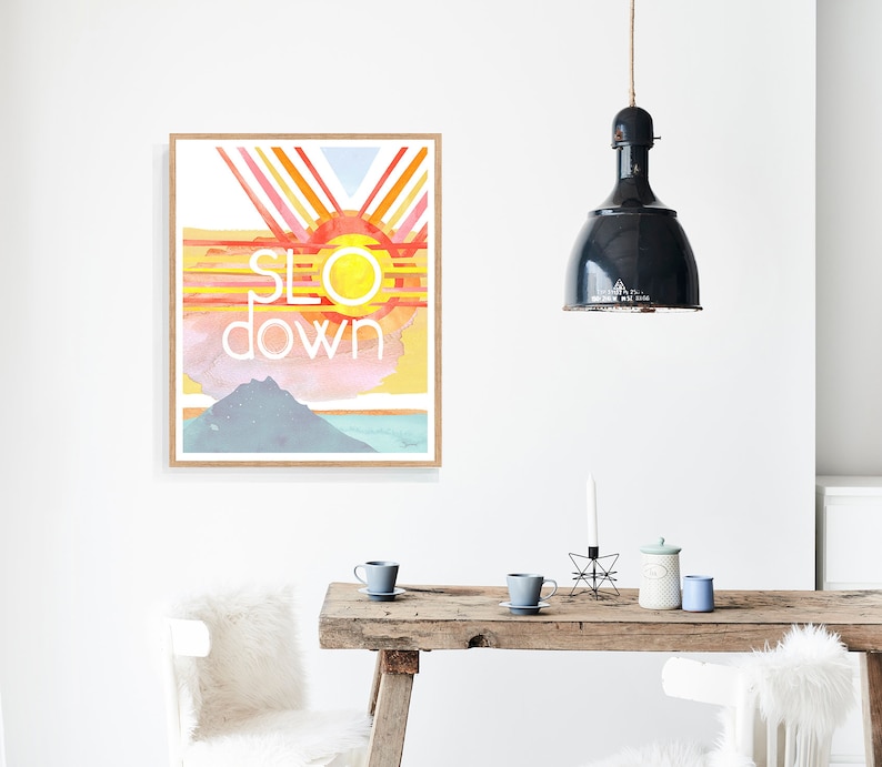 May include: A watercolor print with a sunset scene and the words "slow down" in white text. The print is framed in a light wood frame and is hanging on a white wall.