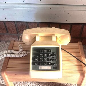 May include: A cream-colored GTE landline telephone with a matching coiled cord and a handset resting on the base. The device features a dark gray keypad with standard numeric buttons, a redial button, a hold button, and a small label area.