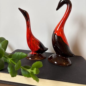 May include: Two ceramic bird figurines with a glossy finish, featuring a vibrant red and deep brown colour gradient. The birds are depicted in a standing position with elongated necks and curved bodies, each mounted on a circular base.