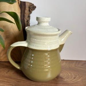Può includere: Una teiera in ceramica dal design bicolore, caratterizzata da una parte superiore e un coperchio color crema maculato che sfumano in una base verde oliva tinta unita. La teiera presenta un manico curvo, un beccuccio corto e una texture rigata.