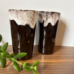 May include: Two ceramic tumblers featuring a glossy dark brown base and a textured, cream-colored drip glaze pattern around the upper rim. The cylindrical vessels have a smooth finish on the lower portion and a bubbly, uneven glaze at the top.