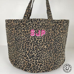 Könnte beinhalten: Eine Tragetasche mit einem braun-schwarzen Leopardenmuster. Die Tasche hat zwei Schulterriemen und die Buchstaben SJP sind in rosa Garn auf der Vorderseite eingestickt. Ein kleines kreisförmiges Logo mit dem Text Lucky Supplies Co ist sichtbar.
