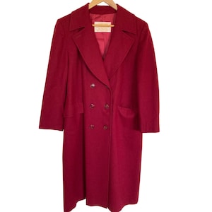 May include: A long, deep red double-breasted coat featuring a wide notched collar, long sleeves, and six dark buttons arranged in two vertical rows. The interior is lined with matching red fabric and includes a rectangular white brand label at the neck.