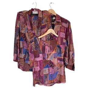 May include: A matching set consisting of a long-sleeve button-up jacket and a short-sleeve top, both featuring a multi-colored geometric patchwork pattern in shades of purple, pink, blue, and brown. The garments are displayed on wooden hangers.