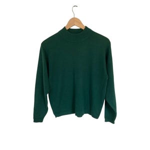 May include: A solid dark forest green long-sleeve sweater featuring a ribbed mock neck collar, ribbed cuffs, and a ribbed hem. The sweater is constructed from a knit fabric and is displayed on a light-colored wooden clothes hanger.