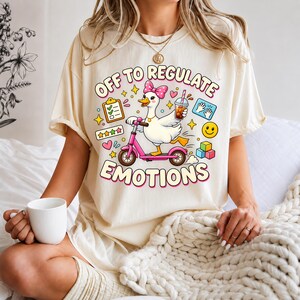 Può includere: Una t-shirt color crema con la grafica di un'anatra bianca con un fiocco rosa a pois, che guida un monopattino rosa tenendo in mano un caffè freddo. Il testo recita OFF TO REGULATE EMOTIONS, circondato da icone di una lista, stelle e cubi.