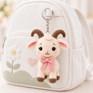 May include: A cream-coloured crocheted goat keyring with brown horns and hooves, featuring a pink bow around its neck, attached to a silver metal ring. The goat is displayed hanging from a white backpack decorated with a white flower and heart.