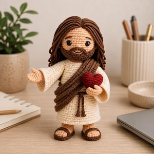 May include: A crocheted doll depicting a figure with long brown hair and a beard, wearing a cream-colored robe with a brown sash and a tie belt. The figure holds a small red crocheted heart in one hand and has the other hand extended outward.