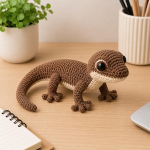 Pode incluir: Uma estatueta de lagartixa feita em crochê nas cores marrom e creme, com superfície de fio texturizado, quatro patas pequenas com dedos distintos, uma cauda longa e curva e grandes olhos redondos de plástico preto brilhante e realista.