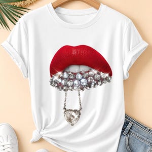 May include: A white short-sleeved t-shirt featuring a graphic print of red lips. The lower lip is adorned with numerous clear, sparkling rhinestones and a silver-toned chain hanging down, which holds a large, faceted heart-shaped crystal pendant.
