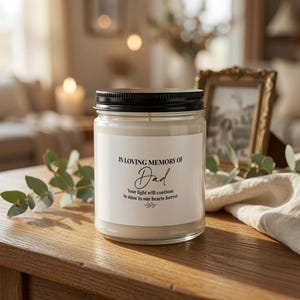 May include: A clear glass candle jar with a black metal lid and a white label. The label features the black text: IN LOVING MEMORY OF Dad, Your light will continue to shine in our hearts forever, with a small leaf graphic below the text.
