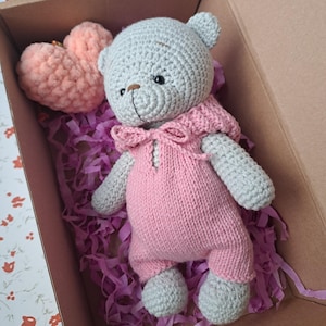 May include: A light gray crocheted teddy bear wearing a pink knitted romper with a bow at the neckline. Beside the bear is a plush, textured peach-colored heart. The items are nestled inside a cardboard box filled with purple paper shred filler.