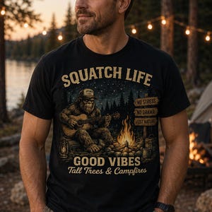 May include: A black t-shirt featuring a graphic of a Sasquatch playing an acoustic guitar by a campfire. The text reads SQUATCH LIFE, NO STRESS, NO DRAMA, JUST NATURE, GOOD VIBES, and Tall Trees & Campfires. The design includes a lantern and signpost.