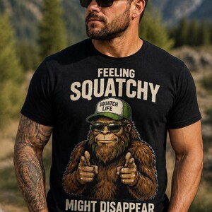 May include: A black short-sleeved t-shirt featuring a graphic of a brown Bigfoot wearing a green cap and sunglasses, pointing forward. The text reads: FEELING SQUATCHY, SQUATCH LIFE, MIGHT DISAPPEAR LATER... IDK. The design is centered on the chest.