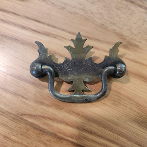 May include: A tarnished brass drawer pull featuring a decorative backplate with a central leaf-like motif and symmetrical side accents. The hardware includes a curved, swinging bail handle attached to two rounded mounting points on the backplate.